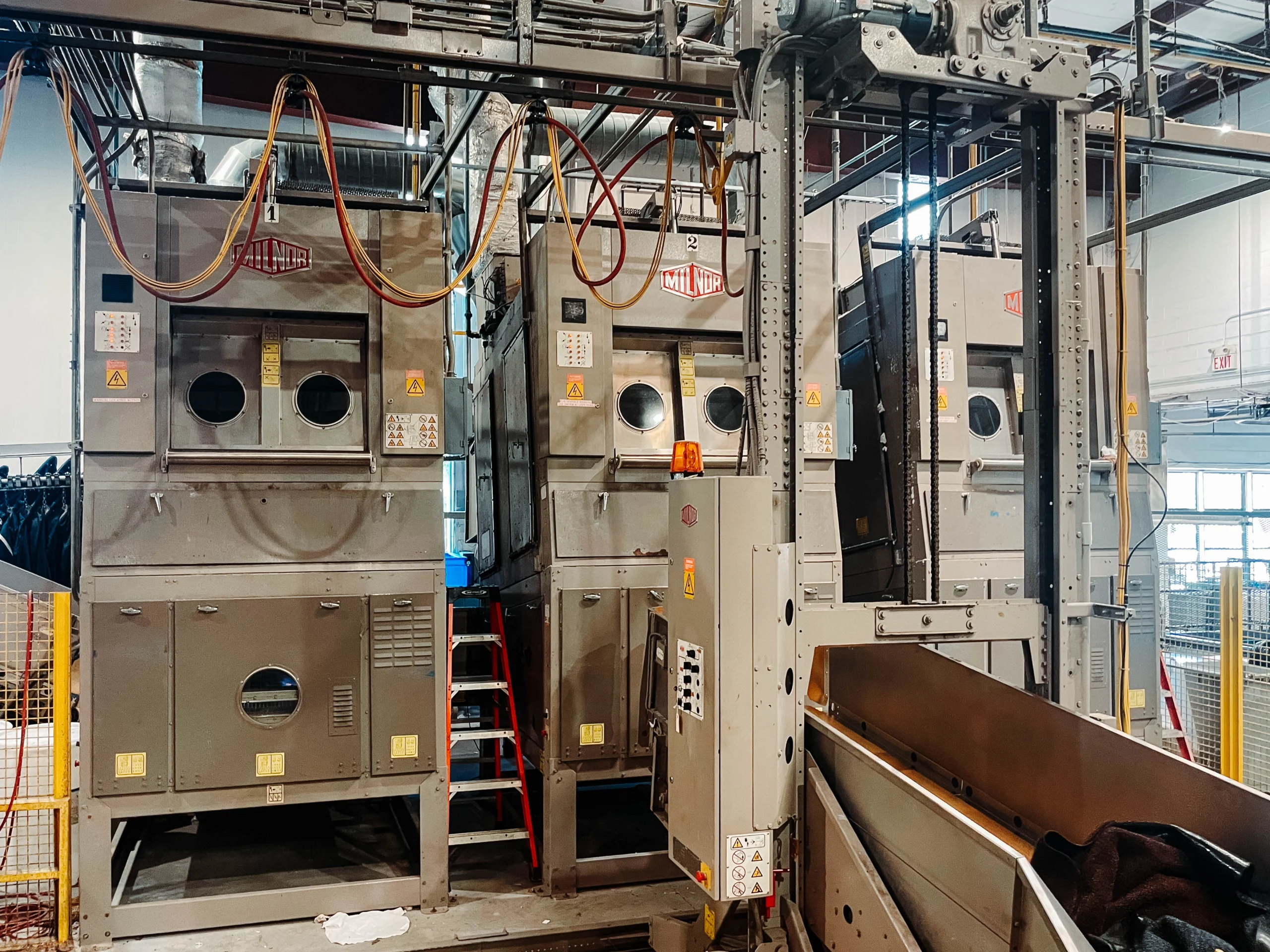 Industrial laundry equipment setup featuring large Milnor washing and drying machines with control panels, piping, and overhead rail systems in a commercial linen facility.