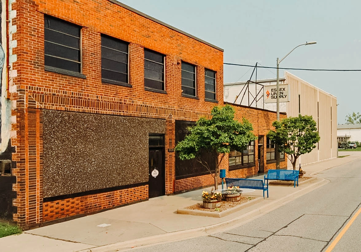 Street view of Essex Linen Supply, featuring a red brick building with large windows, a sidewalk lined with two small trees, flower planters, and bright blue benches. A vintage-style sign hangs on the beige warehouse extension in the background.