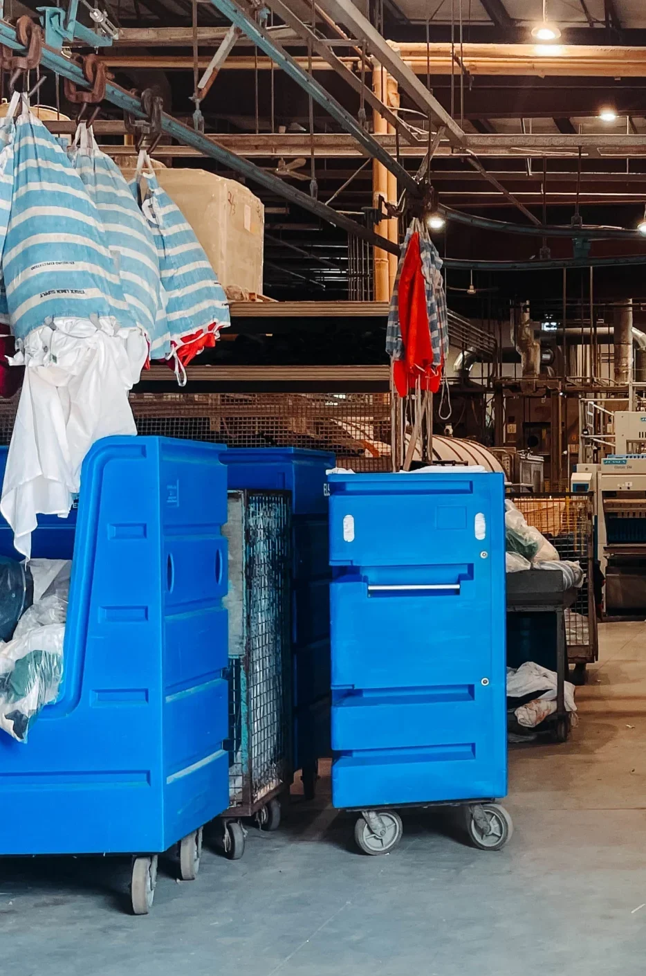 Interior of a commercial linen facility with multiple large rolling carts in blue, green, and beige, filled with linens and towels, surrounded by shelves, hanging garments, and industrial equipment under overhead lighting.