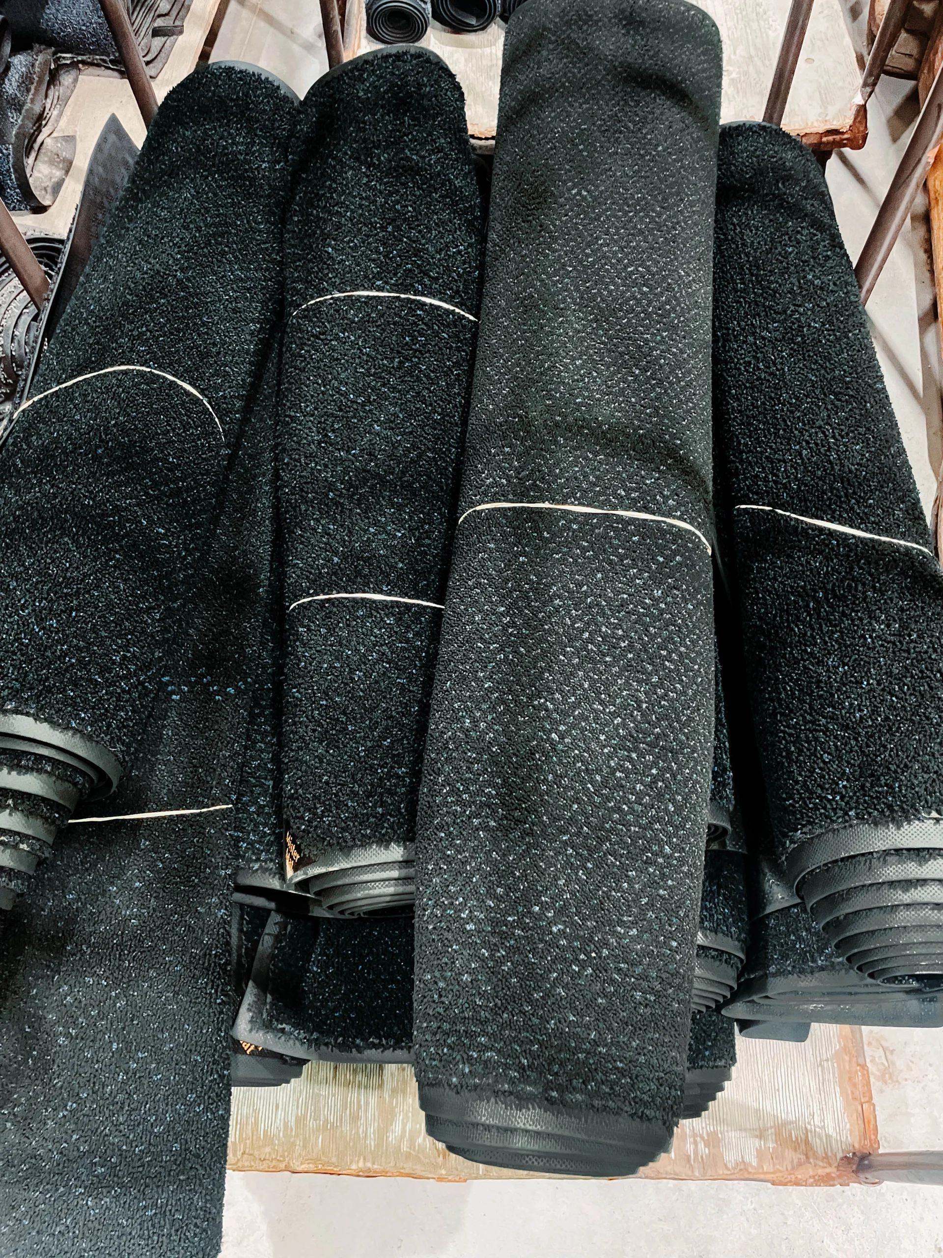 Stack of rolled black commercial floor mats with textured surfaces, secured with bands, arranged on a wooden cart in a linen supply facility.