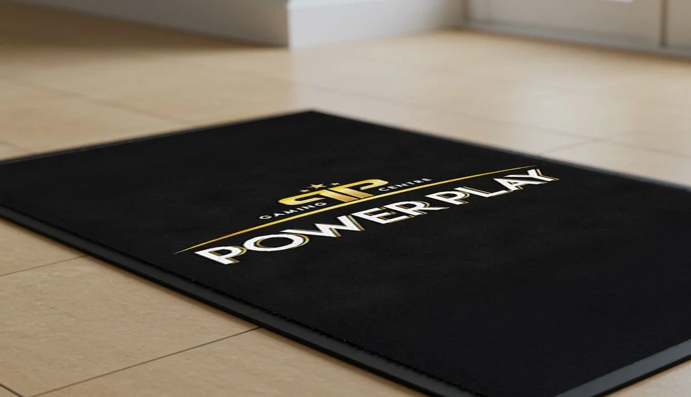 Black floor mat with gold and white text reading “Power Play” and “CIP Gaming Centre,” placed on a light beige tiled floor near large windows in a modern space.
