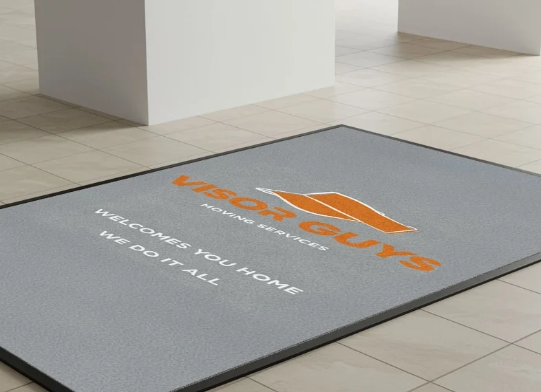 Light grey commercial floor mat with the orange and white logo of Visor Guys Moving Services, featuring the slogan “Welcomes You Home, We Do It All,” placed on a beige tiled floor in a modern building.