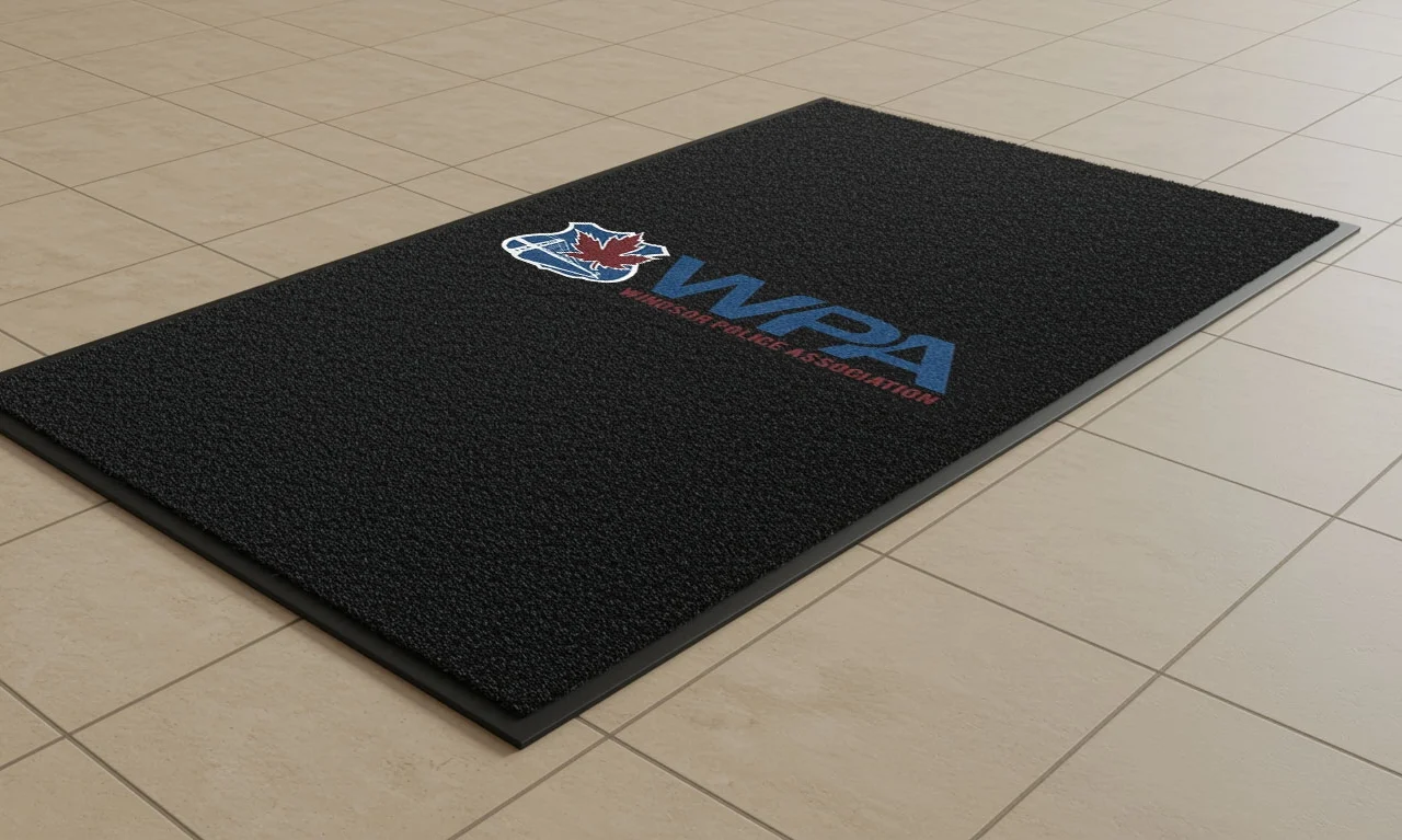 Black commercial floor mat featuring the WPA logo with a red maple leaf and blue text reading “Windsor Police Association,” placed on a beige tiled floor near a wall.