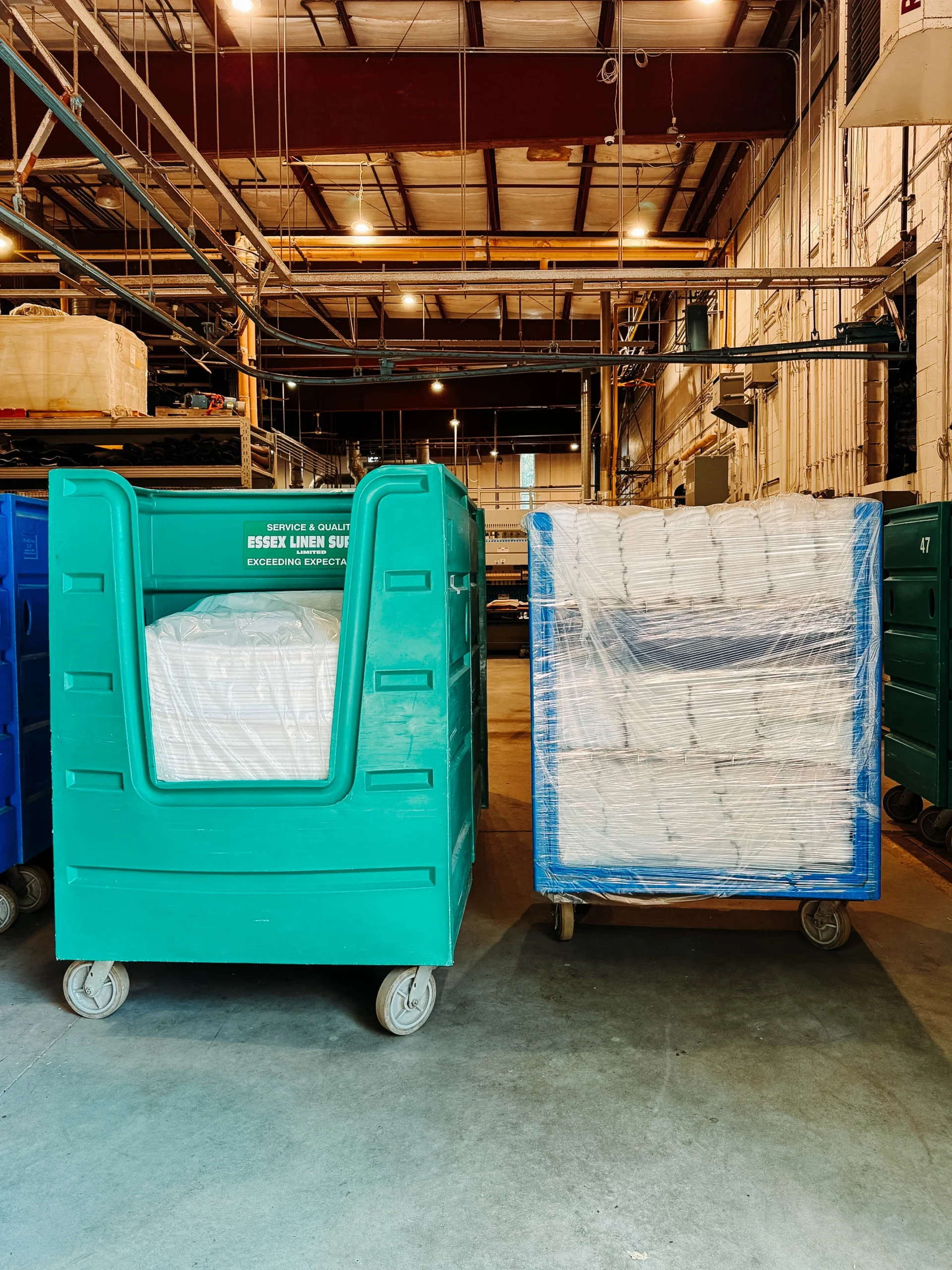 Two large industrial linen carts wrapped in plastic inside a warehouse with high ceilings and exposed piping