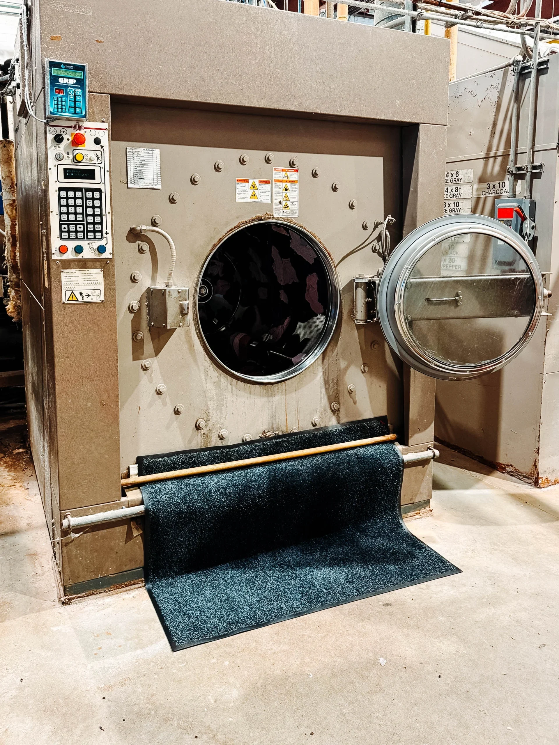 Large industrial dryer with open door and a dark commercial floor mat partially inserted